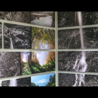 Close, angled view of black-and-white waterfall photographs in the installation, water cascading over dark rock faces beside a color image of backlit autumn leaves over the river.