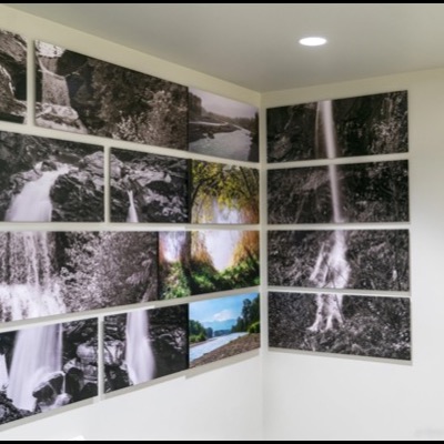 Photograph looking up a stairwell corner at rows of black-and-white waterfall and canyon images alongside a color river photograph, ceiling and recessed lighting visible above.