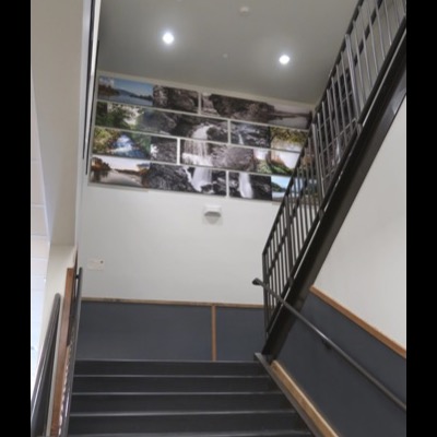 Photograph of the full installation from the base of a stairwell, showing all four rows of river and waterfall panels on the wall above, with the stairs and handrail in the foreground.