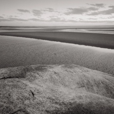 Black-and-white photograph of the boulder’s barnacle-crusted face and vertical fissure, its shadow falling across rippled sand toward the camera.