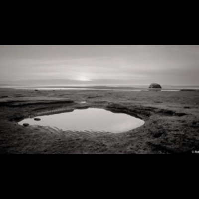 Black-and-white photograph of the split boulder standing alone on an open tidal flat, gentle sand ripples spreading around its base.