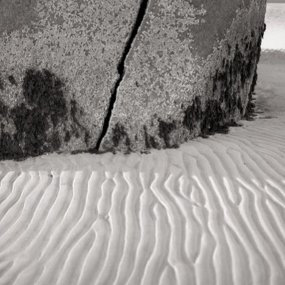 Black-and-white close-up photograph of lichen-speckled rock and its dark central crack, rippled sand meeting the boulder’s barnacled base.