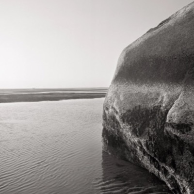 Black-and-white photograph of the boulder’s split profile against the sky, calm water and marsh grass visible along the shoreline.