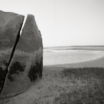 Black-and-white photograph, aerial-feeling, of the boulder viewed from ground level against wide, striped tidal flats reflecting a hazy sky.