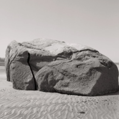 Black-and-white photograph of the full boulder, cracked and barnacle-crusted, resting on rippled sand with a coastline visible in the far distance.