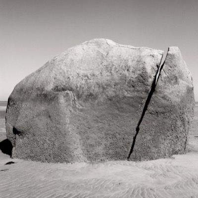 Black-and-white photograph of the boulder’s barnacle-covered shoulder catching low sunlight beside a rocky outcrop, calm water beyond.
