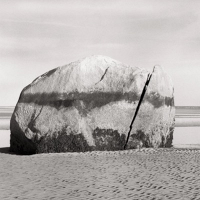 Black-and-white photograph of the split boulder glowing in low evening light, wide tidal flats stretching into the distance.