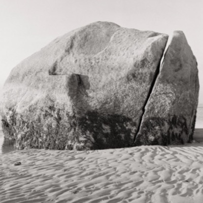 Black-and-white photograph of the split boulder’s two halves side by side, rippled sand and open water surrounding its base.