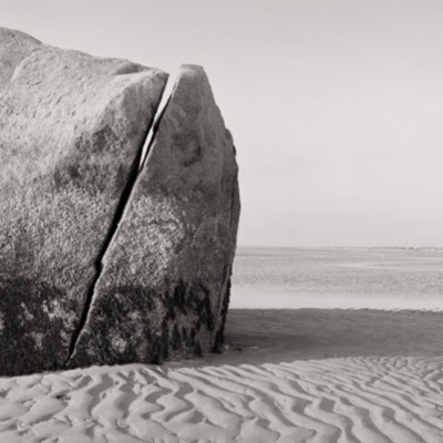 Black-and-white photograph of the boulder standing at the water’s edge, sand ripples in the foreground giving way to distant flats.