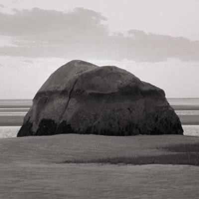 Black-and-white photograph of the boulder under a cloud-streaked sky, its rounded profile catching soft evening light on the flats.