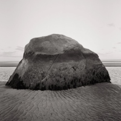 Black-and-white photograph of the boulder’s barnacle-covered lower half against rippled sand, evening light softening its silhouette.
