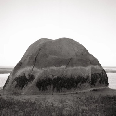 Black-and-white photograph at sunset of open sand ripples and a distant sliver of the boulder’s crack, the sun a bright point on the horizon.