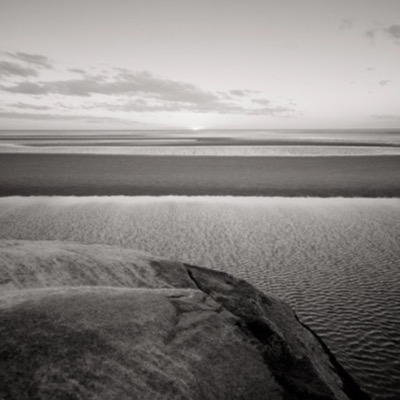 Black-and-white panoramic photograph of the boulder sitting far offshore on the tidal flats, a shoreline of houses and trees stretching along the horizon.