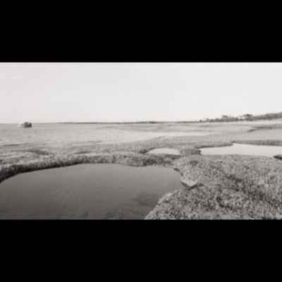 Black-and-white panoramic photograph of a rock jetty stretching into the water at dusk, a small fishing boat idling beside it and beachgoers gathered along the shore.