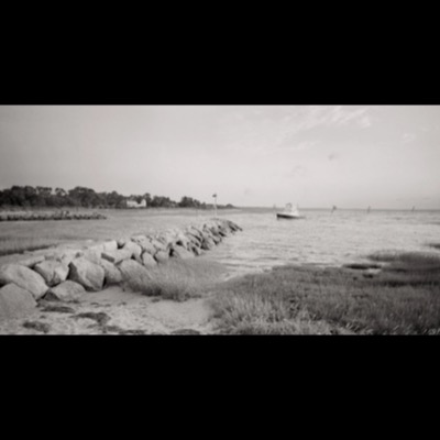 Black-and-white panoramic photograph of a rock jetty stretching into the water at dusk, a small fishing boat idling beside it and beachgoers gathered along the shore.