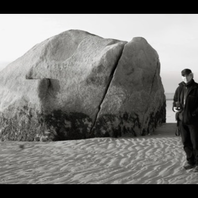 Black-and-white self-portrait of Peter de Lory standing beside the rock on the sand, holding his Hasselblad camera, the tidal flats stretching behind him.